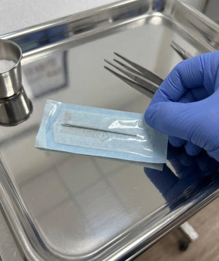 A gloved hand holds a packaged needle above a metal preparation tray