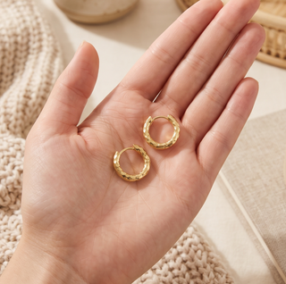 Textured gold hoop earrings with a hammered finish and polished golden shine, displayed in an open palm against a soft, neutral fabric background.