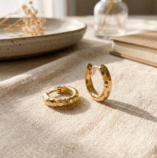 A pair of chunky gold hoop earrings featuring a distinctive hammered texture and hinged closure, styled on beige linen with warm sunlight for a timeless, everyday jewelry look.
