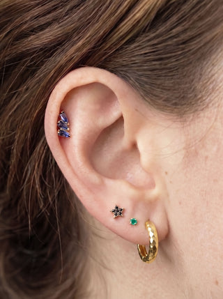 Gold hammered hoop earring paired with a green gemstone stud, black star-shaped stud, and blue gemstone cartilage jewelry, showcasing a curated ear stack with mixed colors and textures.