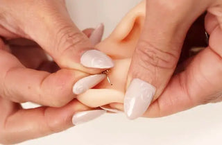 A delicate gold-colored stud earring being carefully placed into the ear of a doll with pale skin.