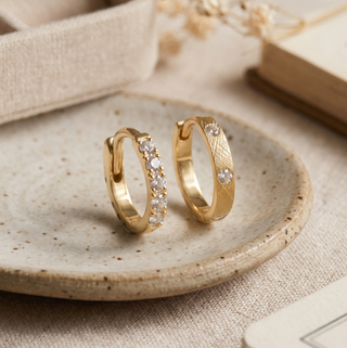 Two gold huggie hoop earrings with sparkling diamond accents, featuring a classic pavé design and a textured crosshatch finish, styled upright on a neutral ceramic dish.