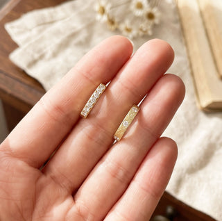 Close-up of gold diamond huggie earrings showcasing a pavé-set design on one pair and a crosshatched gold finish with scattered diamond accents on the other, displayed on fingertips in natural light.