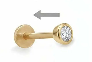 Gold-colored labret piercing with a round, clear gemstone set in its front end.