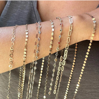 Nina Wynn permanent jewelry inspiration: gold-tone paperclip, disc and stone-accent chain samples draped across a forearm.